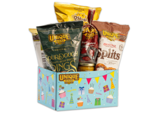 varied unique snacks products in a light blue box with different colored present boxes party hats, and cupcakes all over it with the unique snacks logo in the upper left hand corner